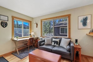 Un salon chaleureux avec un canapé en cuir, un bureau en bois et une fenêtre donnant sur des arbres et des montagnes.