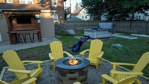 Een achtertuin met een vuurkuil, omringd door gele Adirondack-stoelen, een barbecueplaats en een tuinhuis.