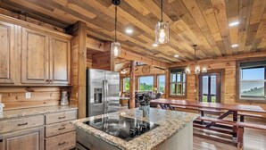 Eine Küche mit Holzschränken, Granit Arbeitsplatten, einem Edelstahlkühlschrank und einer großen Kochinsel mit Induktionskochfeld. Im angrenzenden Bereich befinden sich hölzerne Bänke und ein Esstisch.