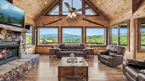 Ein geräumiges Wohnzimmer mit Kamin aus Stein, Ledersofas, einem Holztisch und großen Fenstern mit Blick auf die Berge.