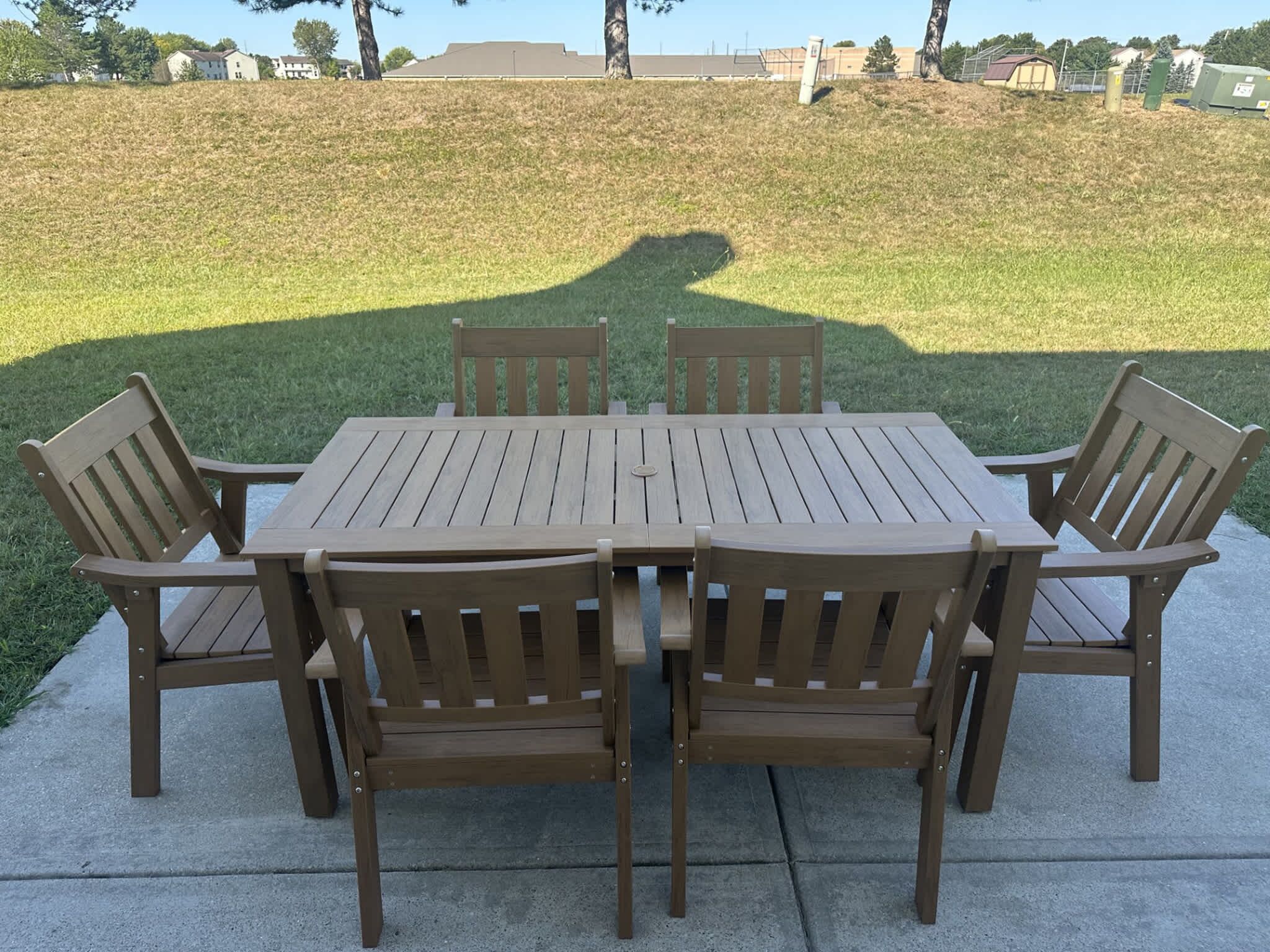 A wooden patio table with six chairs, set on a concrete surface with a grassy hill and buildings in the background.