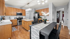 A modern kitchen with wooden cabinets, stainless steel appliances, a dining area with a patterned tablecloth, and a chandelier. - Pickerington Haven Home for 10 (Pickerington)