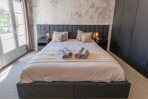 A neatly made bed with two rolled towels, a headboard, bedside tables, and a view of the outdoors through a window.