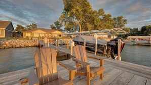 Un muelle de madera con dos sillas Adirondack, un barco y una casa al fondo.