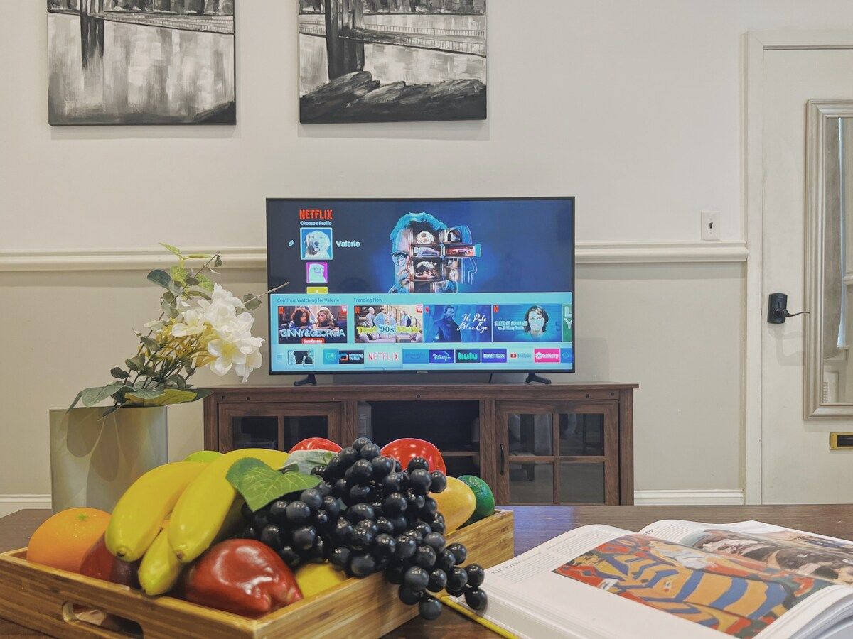A room with a television displaying a Netflix interface, a fruit bowl, and a book on a table.