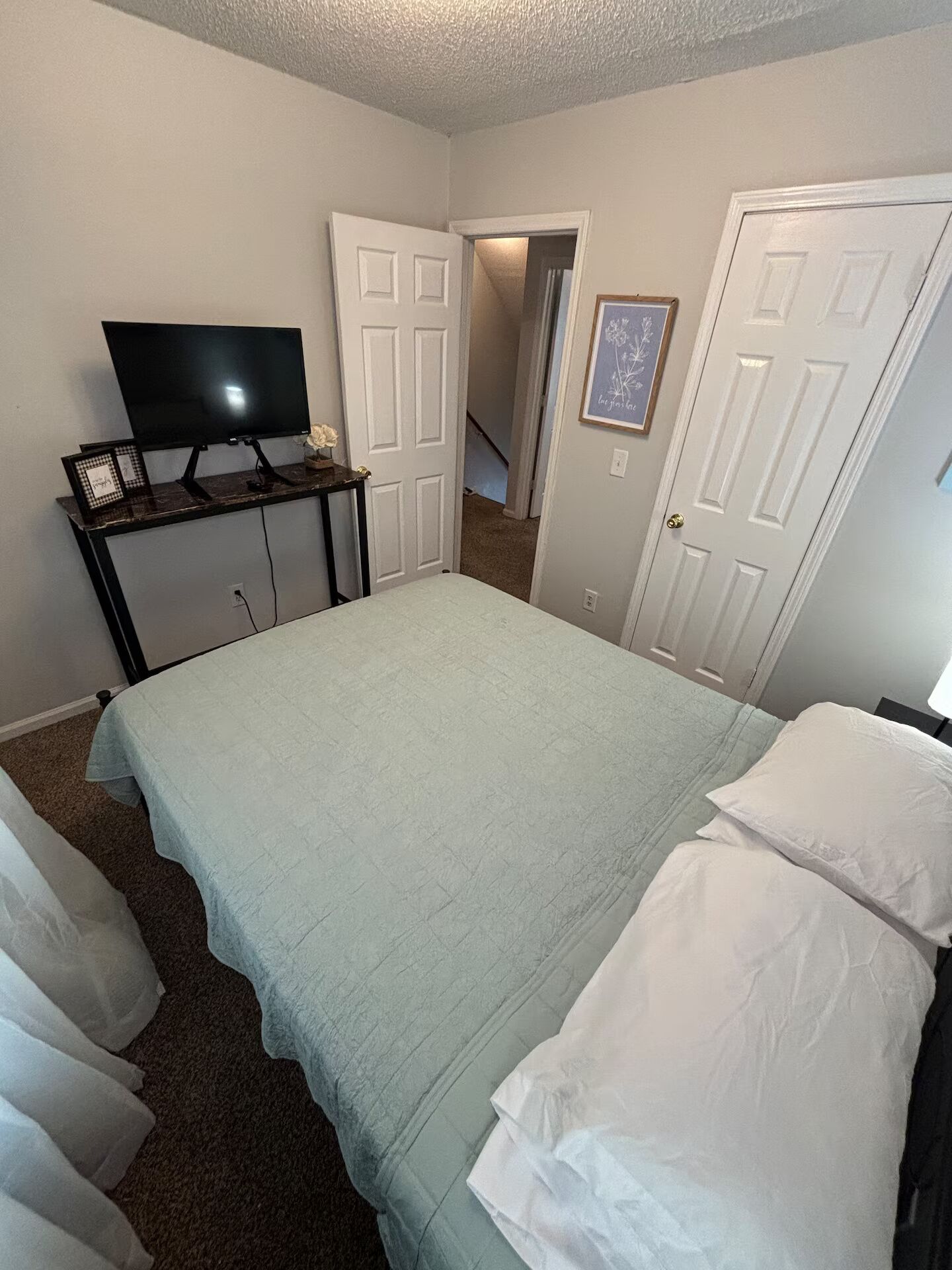 A bedroom with a bed, a TV on a stand, a door, and a hallway.