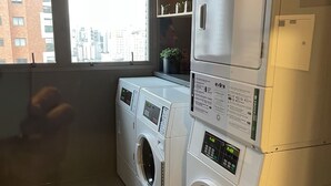 A laundry room with two front-loading washing machines, a small shelf with decorative items, and a window with a city view. - BEAUTIFUL AND COZY STUDIO (São Paulo)