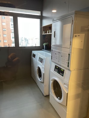 Un cuarto de lavandería con dos lavadoras de carga frontal, un estante pequeño con objetos decorativos y un ventanal con vista a la ciudad.