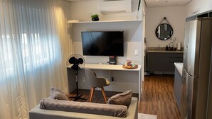A modern living room with a sofa, a desk with a computer, a flat-screen TV, and a kitchen area with a refrigerator and sink. - BEAUTIFUL AND COZY STUDIO (São Paulo)