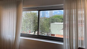 A room with a large window, white curtains, and a view of buildings and trees outside. - BEAUTIFUL AND COZY STUDIO (São Paulo)