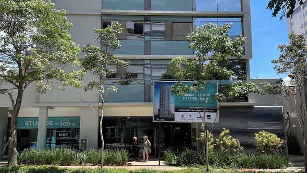 A modern building with a large glass facade, a signboard advertising apartments, and a gated entrance. - BEAUTIFUL AND COZY STUDIO (São Paulo)