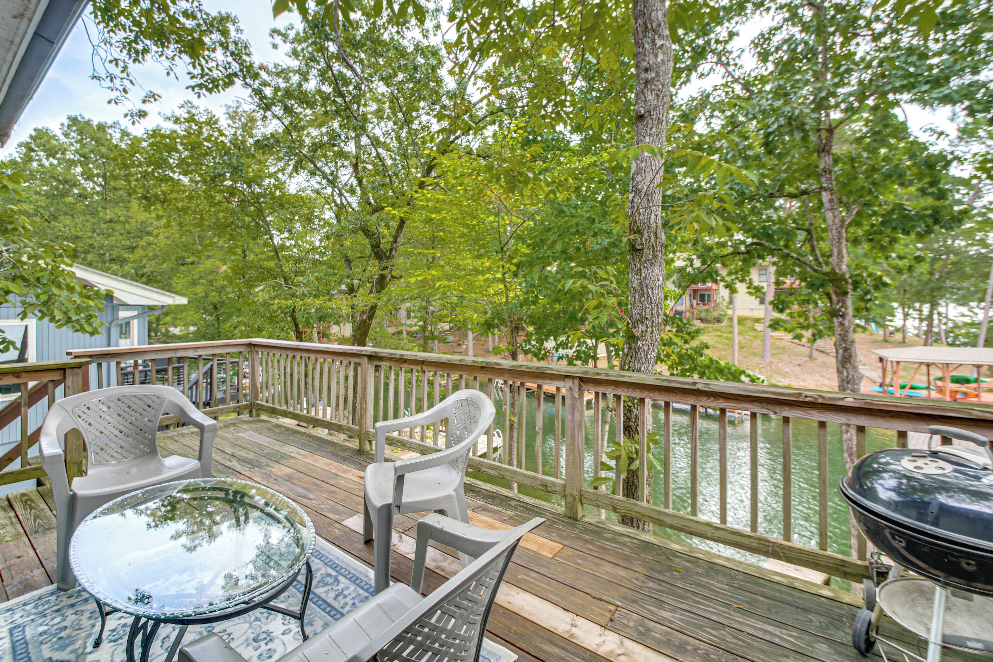 A wooden deck with a glass table and chairs, a grill, and a view of a lake.