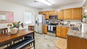 A kitchen with wooden cabinets, stainless steel appliances, and a dining area with a table and chairs. - < 1 Mi to Ole Miss & The Grove! Charming Getaway (Oxford)