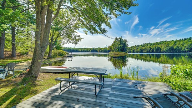 A wooden deck with a picnic table and chairs overlooking a calm lake, surrounded by trees and greenery. - Waterfront Escape w/ Beach Access in Bridgton (Bridgton)