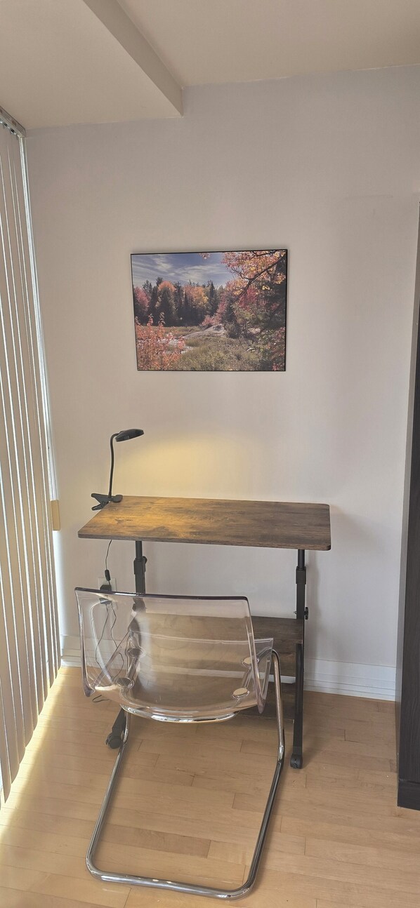 A room with a wooden desk, a clear plastic chair, and a framed picture of a landscape on the wall.