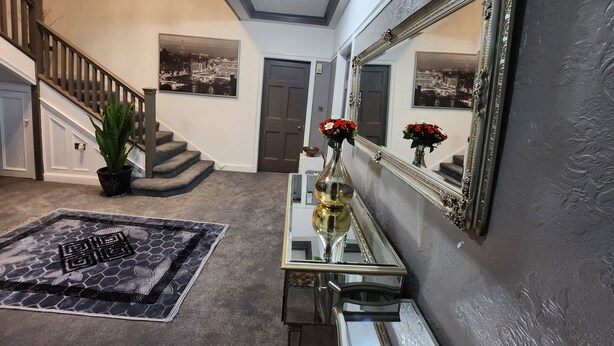 Un couloir avec un escalier, une console sur laquelle est posé un vase de fleurs, et un grand miroir au mur.