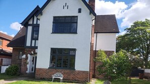 Une maison à deux étages, avec une façade blanche et des éléments décoratifs sombres, un porche à l’avant avec un banc, et une petite pelouse agrémentée d’une table et de chaises.