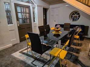 Een eethoek met een glazen tafel, zwarte en gele stoelen, een bank en een klein raam met een schilderij.