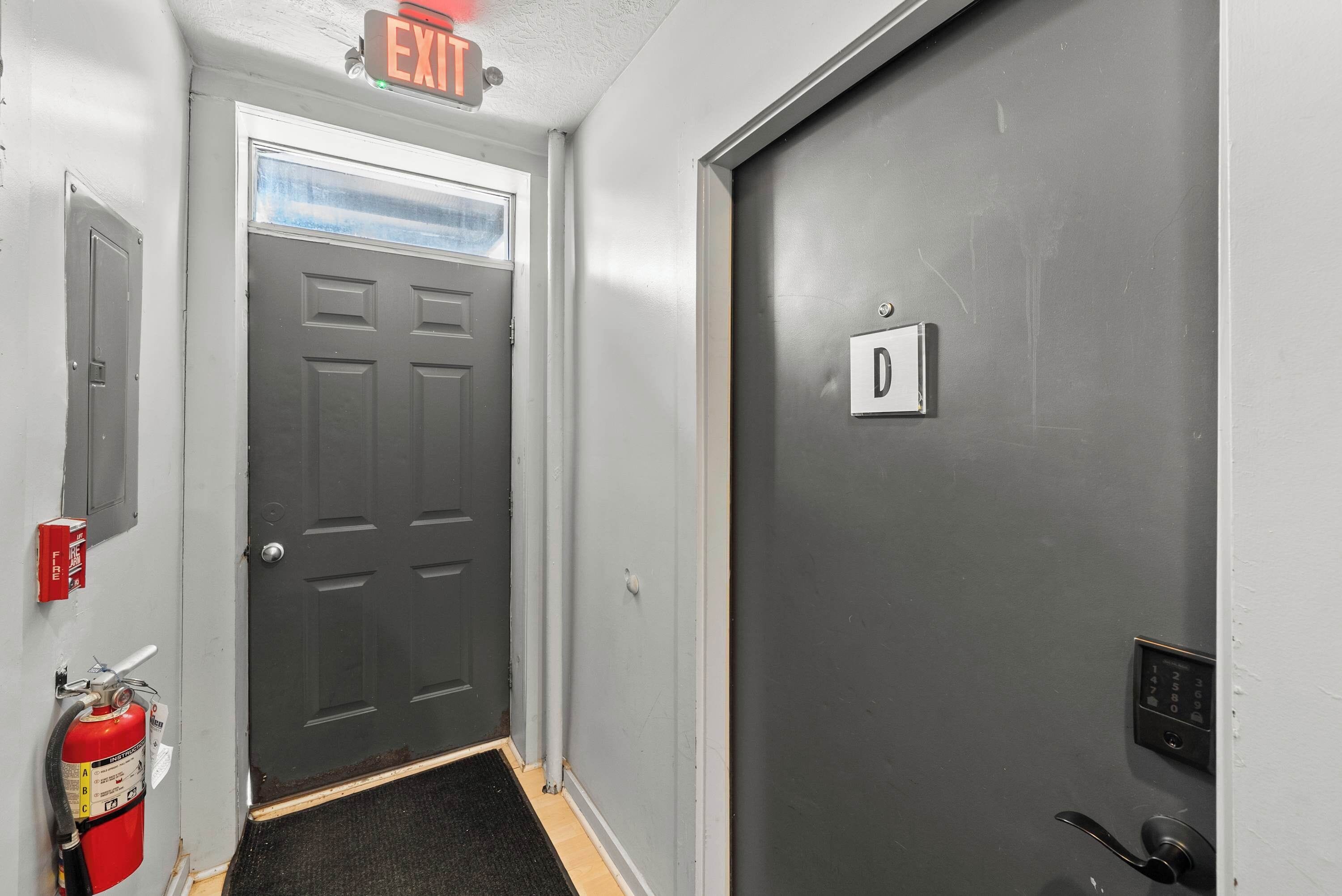 A hallway with a grey door marked 'D', a fire extinguisher, and an exit sign.