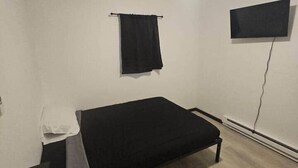 A small, sparsely furnished room with a single bed, a ceiling fan, a flat-screen TV mounted on the wall, and a window with a black curtain. - Happy Trails Hideout #2 (Houston)
