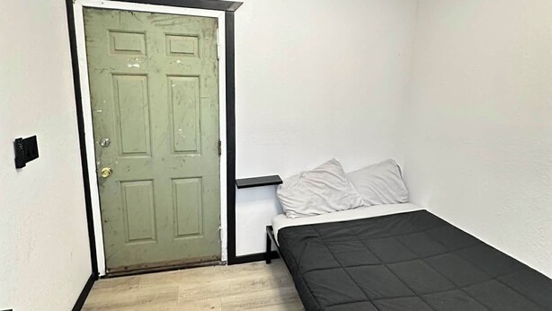 A small, single-room apartment with a green door, a bed with a black mattress, and a small trash bin. - Cozy Camo Cottage #8 (Houston)