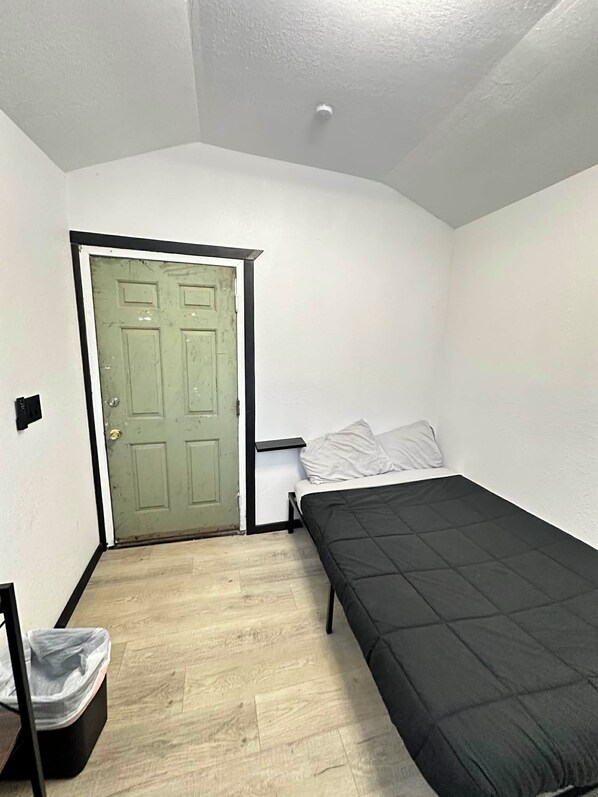 A small, single-room apartment with a green door, a bed with a black mattress, and a small trash bin. - Cozy Camo Cottage #8 (Houston)