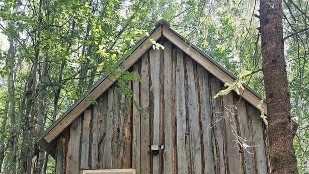A rustic wooden cabin with a white door, surrounded by trees and a forest floor. - Rustic Bunk #5 (Houston)