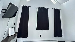 A room with a sloped ceiling, skylights, a bed, a desk, and black curtains. - Rustic Bunk #5 (Houston)