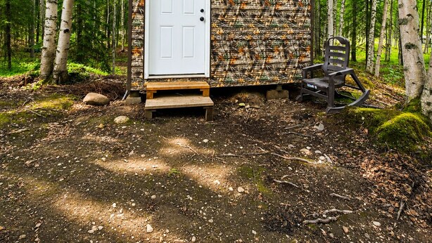 A small cabin with a white door, a bench outside, and a forested backdrop. - Mat-Su Thunder Cabin #1 (Houston)