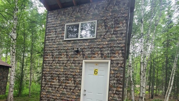 A rustic cabin with a white door marked with the number 6, a red folding chair outside, and a natural forest backdrop. - Vibing In The Woods #6 (Houston)
