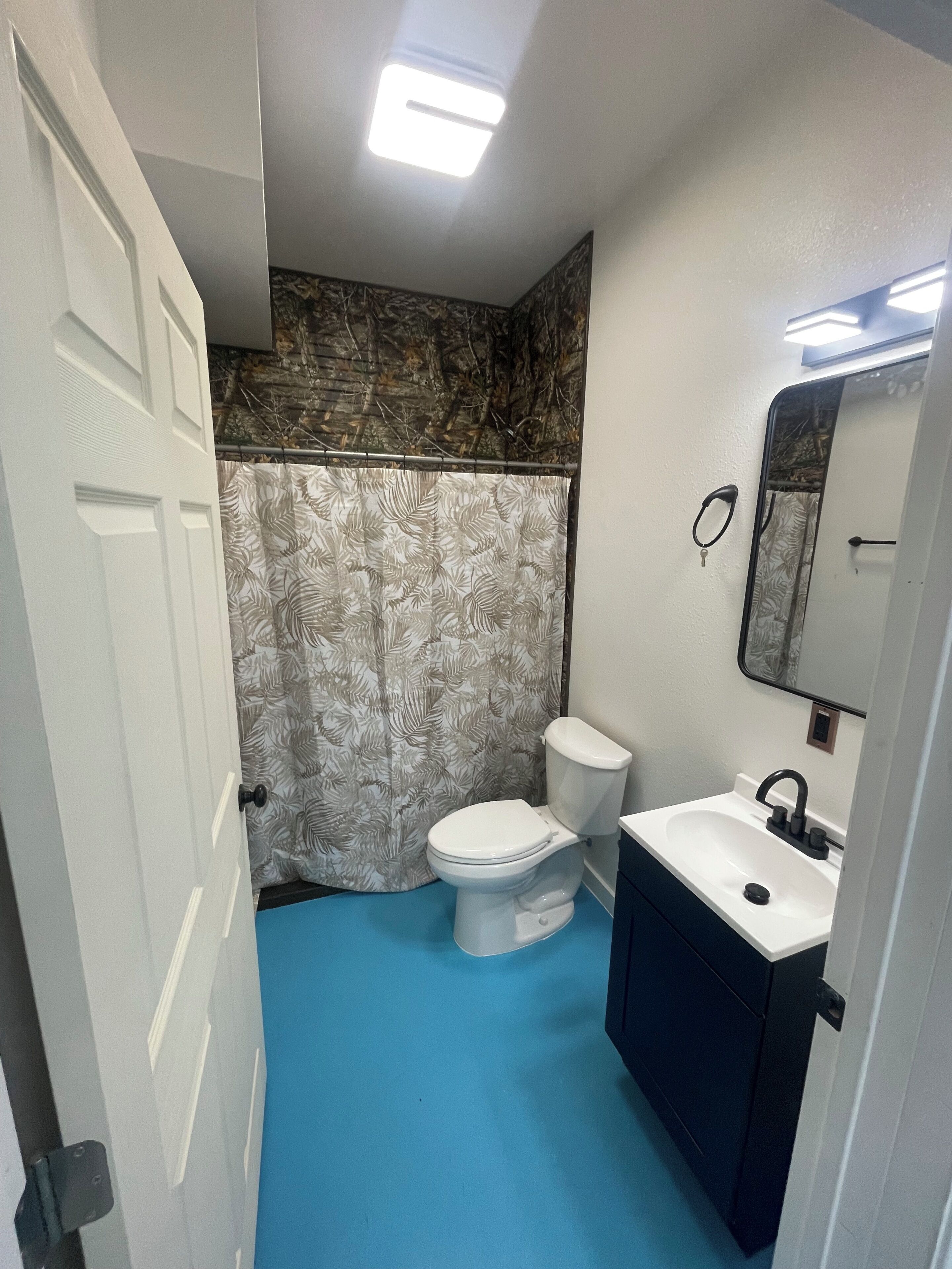 Ein Badezimmer mit einer weißen Toilette, einem Waschbecken mit einem schwarzen Wasserhahn und einer Duschvorhang mit Blättermotiv.