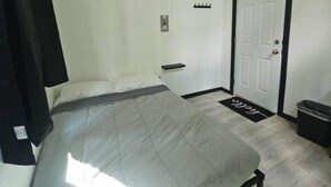 Quarto com cama, ventilador de teto, porta e uma pequena lixeira.