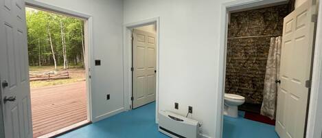 Un pasillo con piso azul, un calentador blanco, una puerta blanca que da a un baño con pared de piedra, y una puerta a un área exterior con sendero de ladrillos y árboles.