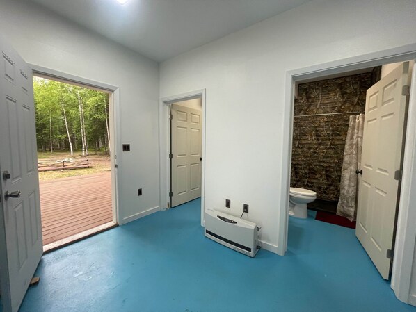 A hallway with a blue floor, a white heater, a white door leading to a bathroom with a stone wall, and a door to an outdoor area with a brick path and trees.
