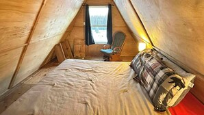 A cozy, triangular wooden tent with a bed, a chair, and a window with curtains. - Bass Farm: Lake View Loft Cabin (Houston)
