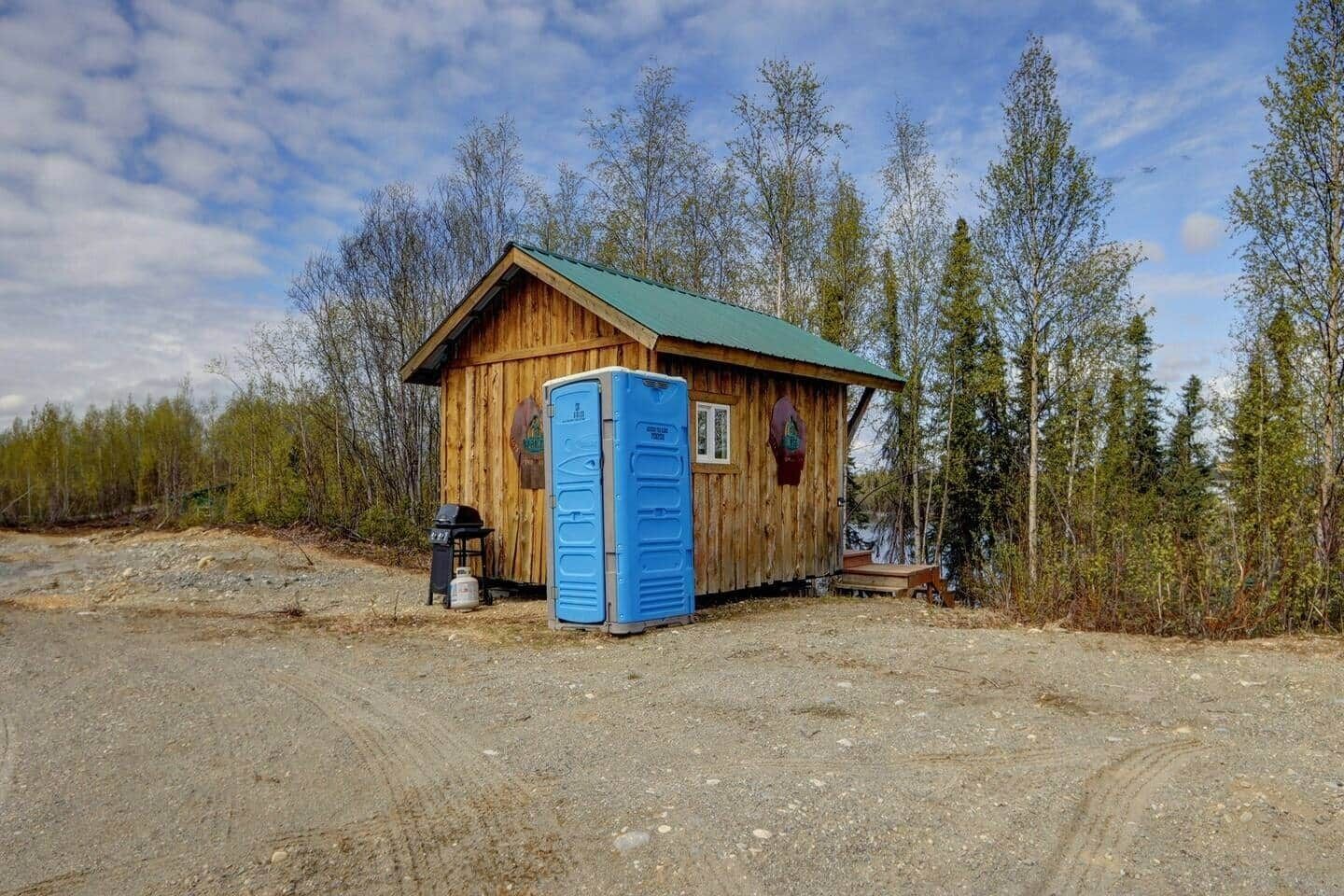 Una cabaña de madera con techo verde, un baño químico azul y una pequeña zona de estar al aire libre.