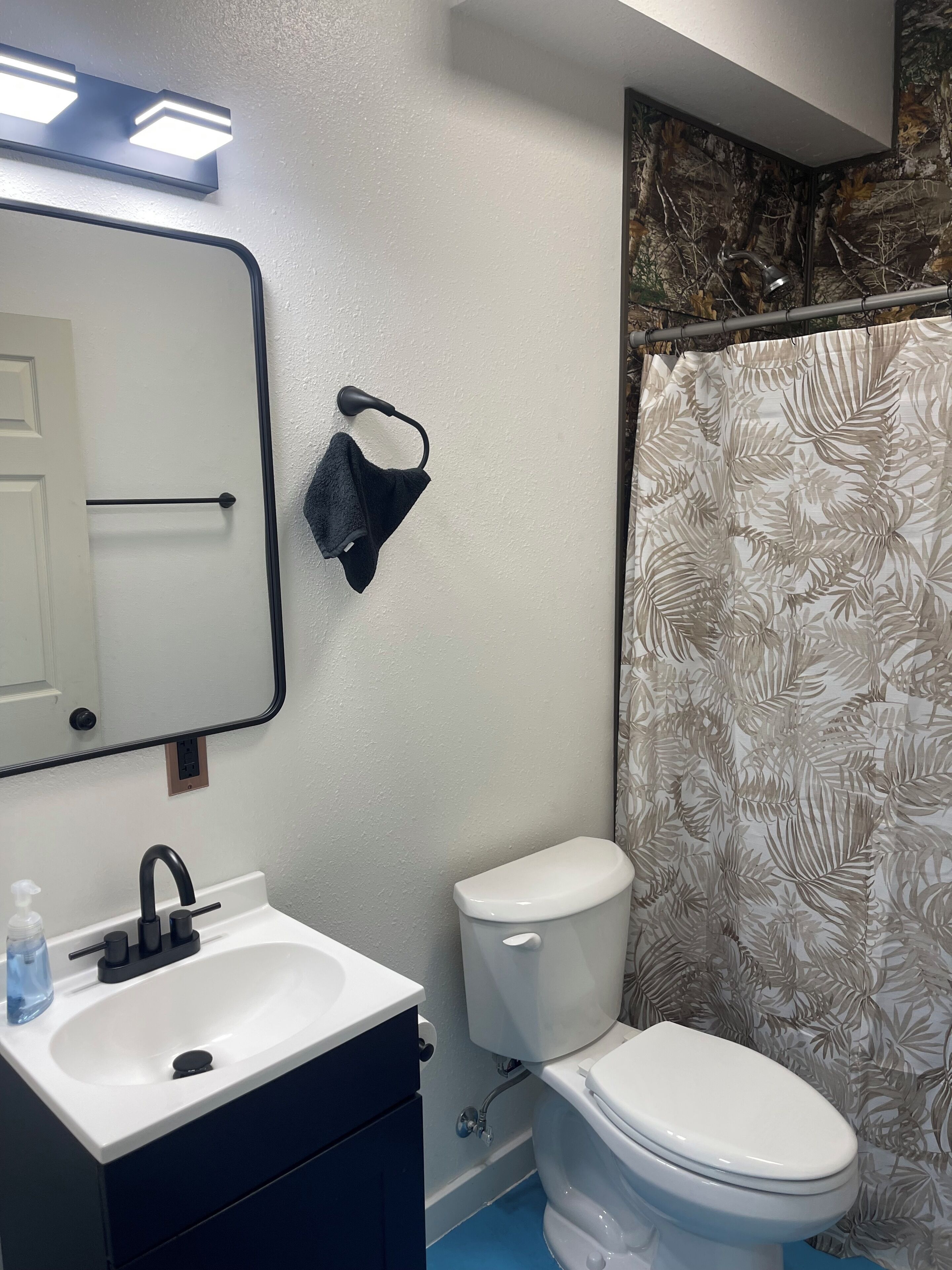 A bathroom with a white sink, black faucet, and a toilet. There is a towel rack with a black towel hanging on it. The walls are painted white, and there is a shower curtain with a leaf pattern.