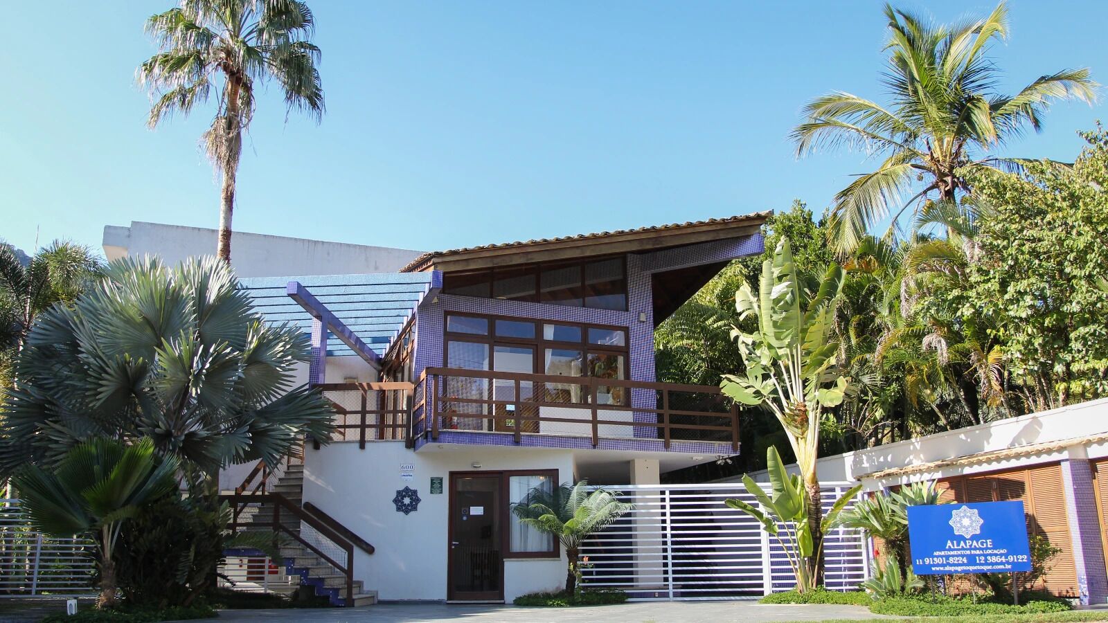 A two-story building with a balcony, surrounded by palm trees and a clear sky.