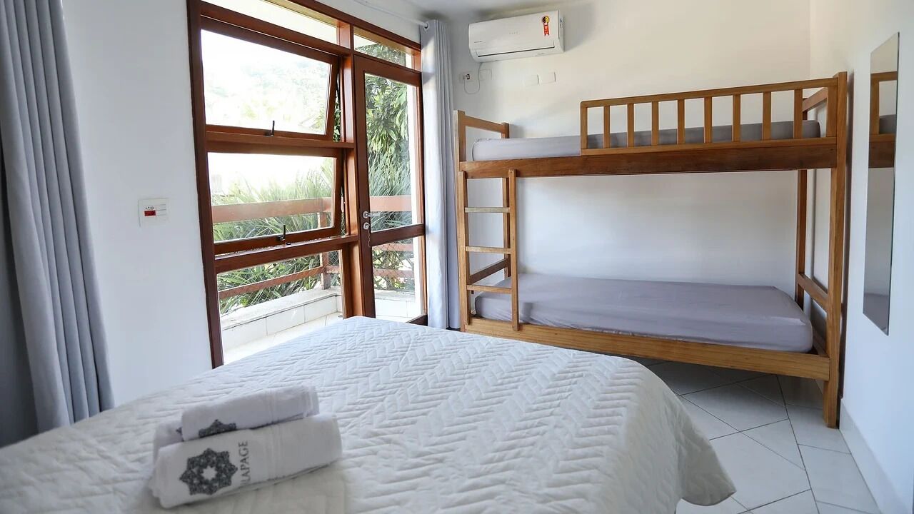 A room with a bunk bed, a bed with a quilt, a window with curtains, and an air conditioning unit on the wall.