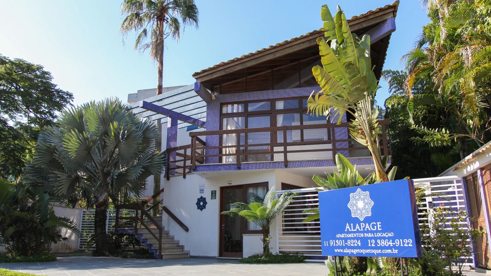 A two-story building with a balcony, surrounded by palm trees and a gated entrance.