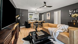Una sala de estar moderna con sofá, mesa de centro y televisor. Se ve una zona de comedor con mesa y sillas al fondo.