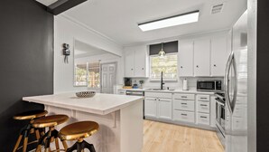 Una cocina moderna con armarios blancos, una isla central con taburetes, electrodomésticos de acero inoxidable y un ventanal con persianas.