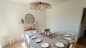 A dining area with a round table set for six, white chairs, a chandelier, a mirror, and a shelf with decorative items.