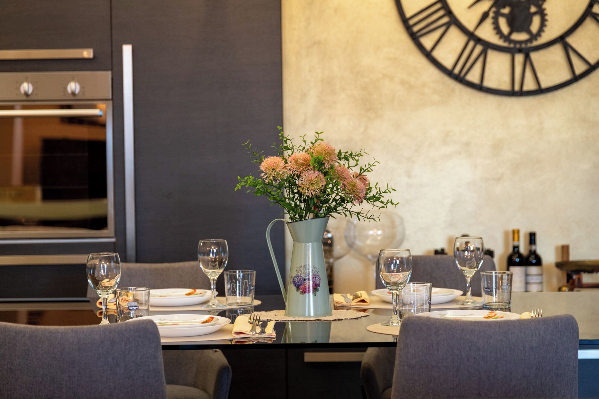 Une table à manger dressée avec des assiettes, des verres et des couverts, un vase rempli de fleurs et une horloge accrochée au mur.