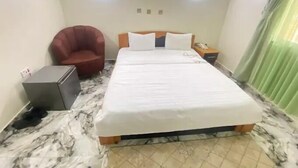 A hotel room with a bed, a chair, a nightstand, and a small table. - Starwave Homes (Abuja)