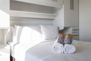 A neatly made bed with white linens and towels, a white wardrobe, and a shelf above the bed. - Code Aclimação (São Paulo)