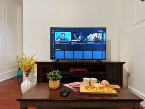 A living room with a flat-screen TV on a wooden stand, a coffee table with a tray of pastries, a remote control, and decorative plants.
