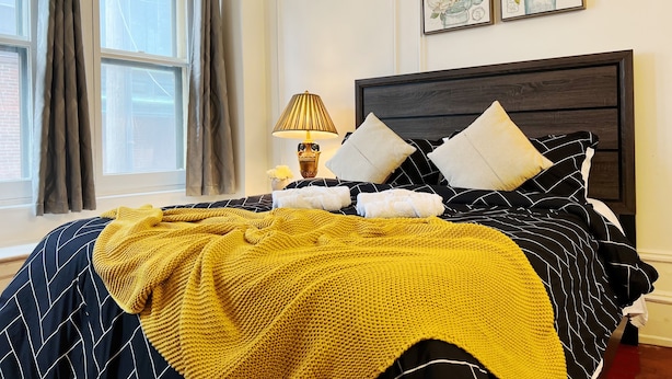 A neatly made bed with a black and white striped bedspread, a yellow knitted throw, and two white pillows. There is a nightstand with a lamp and a vase of flowers beside the bed. Two framed floral artworks are hung on the wall above the bed. - Ramblei near Rittenhouse Sq / 2 bedroom – Unit B (Philadelphia)