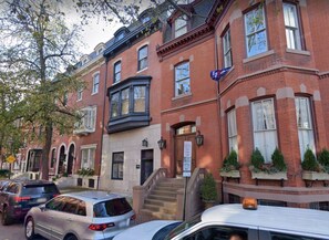 A row of brick townhouses with a central entrance, steps leading to the door, and a small garden area with potted plants.