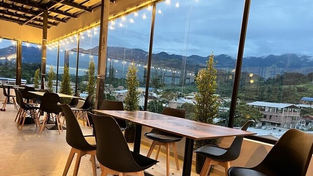 Een eetruimte met tafels en stoelen, grote ramen met uitzicht op bergen en een brug, en lichtslingers.